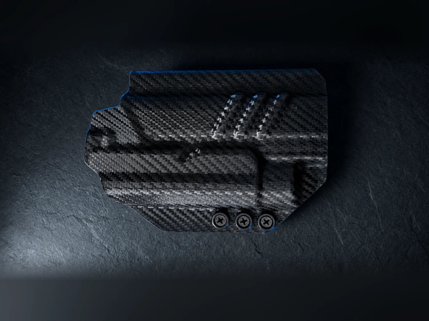 Black carbon-fiber Kydex holster shell on dark slate, molded to the exact contours of a pistol slide, trigger guard and weapon light, edged with a cool blue rim light
